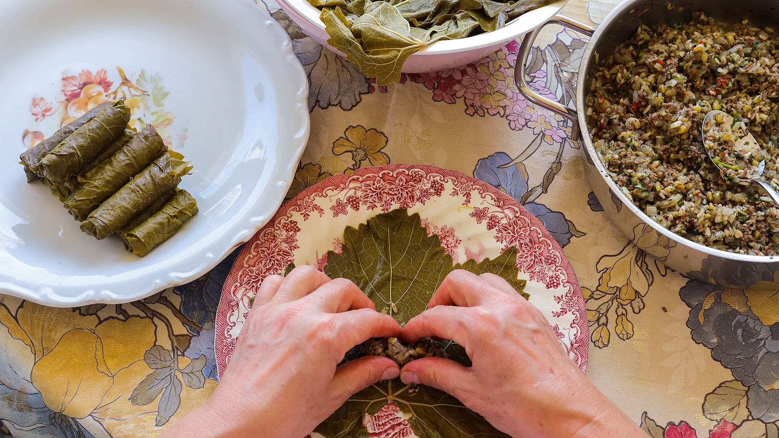 Hands rolling stuffed grape leaves with a mixture, on a floral tablecloth, surrounded by prepared rolls and filling in a pot.