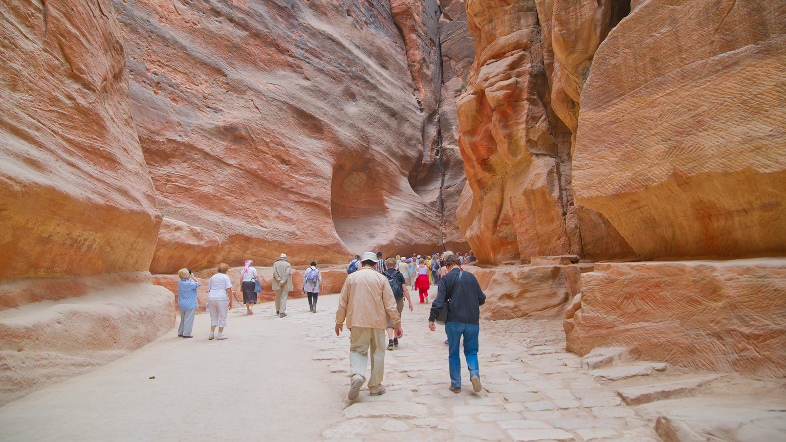 Tourists walking through a narrow, towering canyon with reddish rock walls in Petra, Jordan, under a clear sky.
