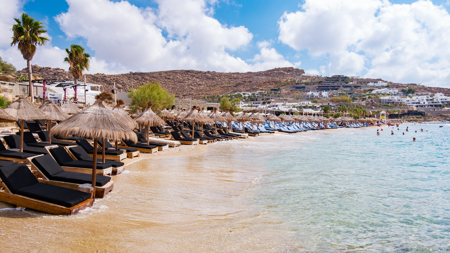 A beach with rows of thatched umbrellas and lounge chairs, calm turquoise water, and a rocky hillside with white buildings in the background.