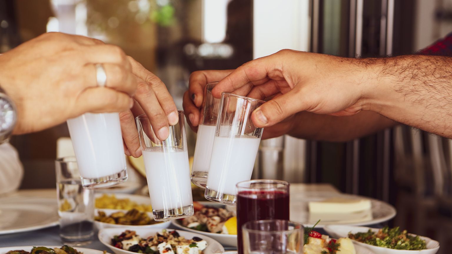 Three hands clinking glasses filled with a milky white beverage over a table with various appetizers.