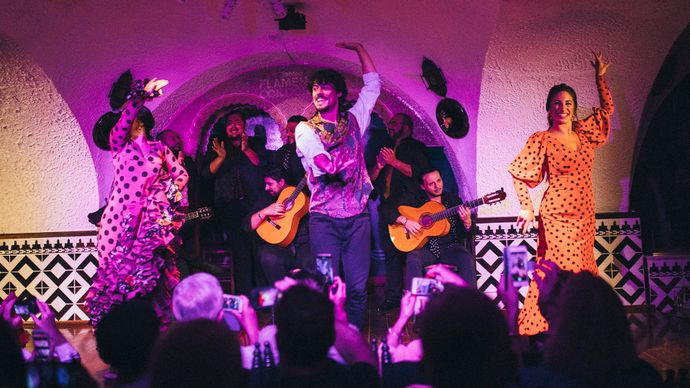 Flamenco dancers in polka dot dresses perform energetically with guitarists on stage, as an audience captures the scene with phones.