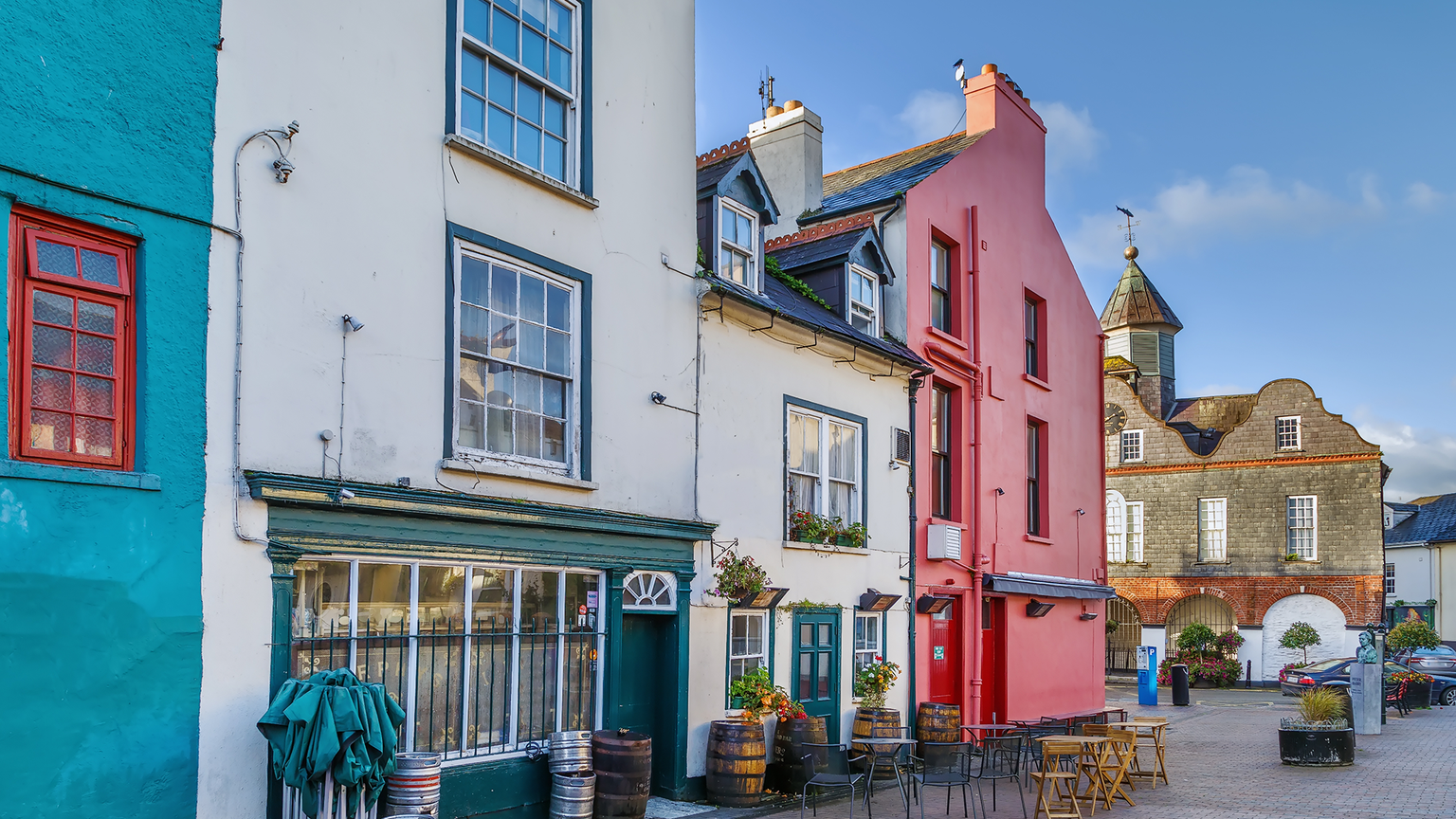 Colorful buildings with green, white, and red facades, a cobblestone street, outdoor tables, and a tower in the background.