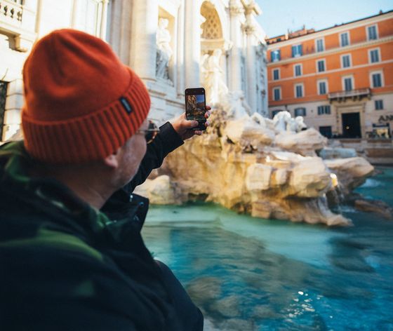 Person in orange beanie taking a selfie with a smartphone near a large ornate fountain in a sunny urban setting.