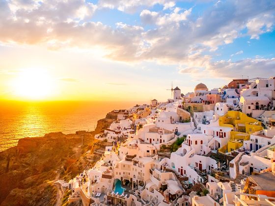 Sunset over Santorini, Greece, with whitewashed buildings and blue domes perched on a hillside, overlooking the Aegean Sea.