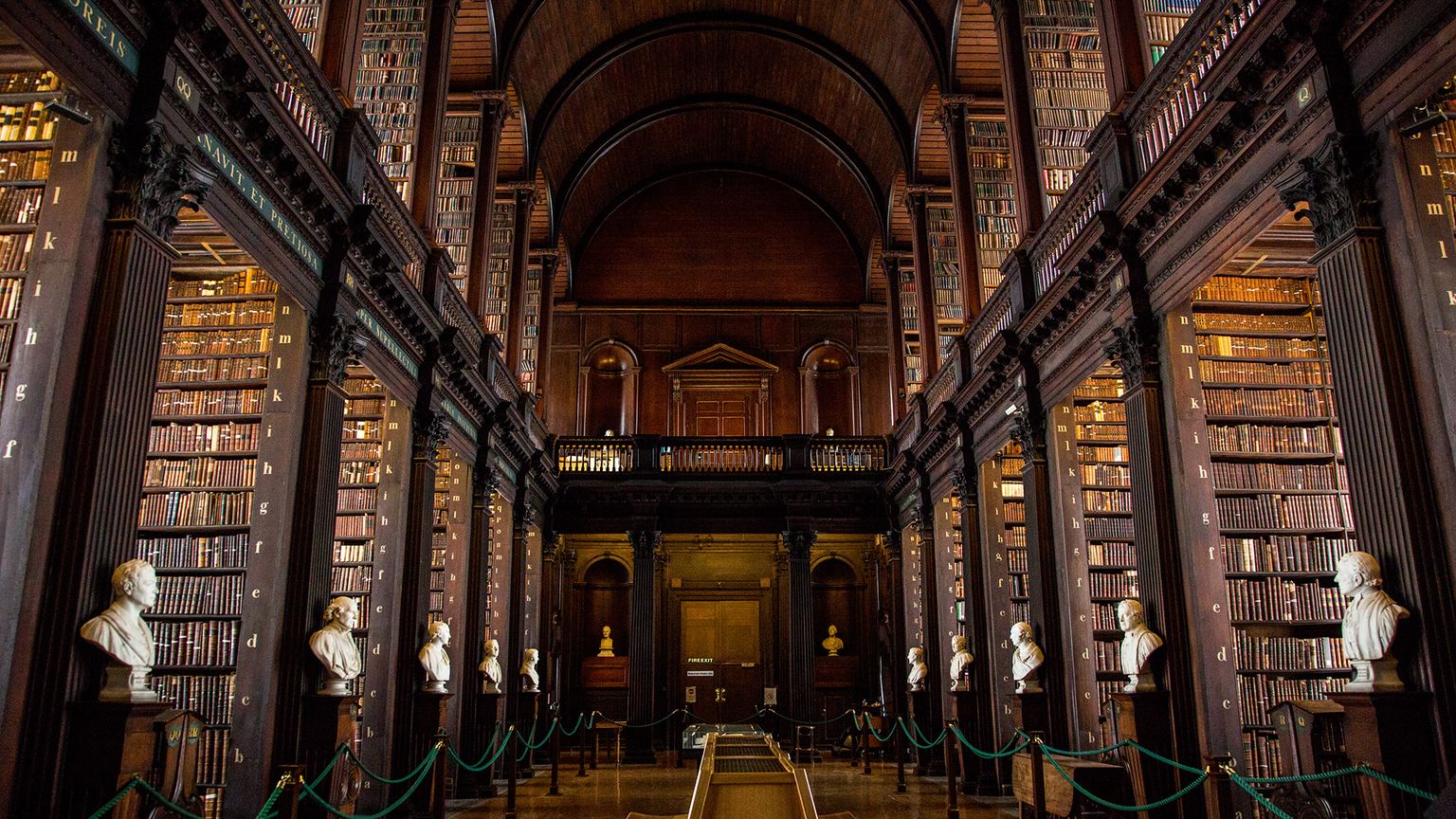 Ornate library with tall wooden shelves filled with books, arched ceiling, and marble busts lining the aisle.