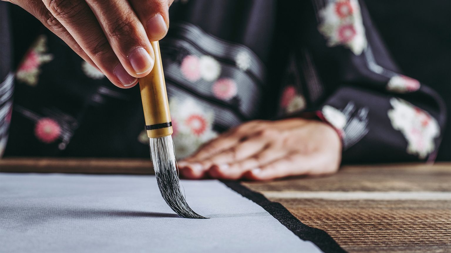 Person in floral-patterned clothing delicately painting on paper with a calligraphy brush, focused on creating precise strokes.