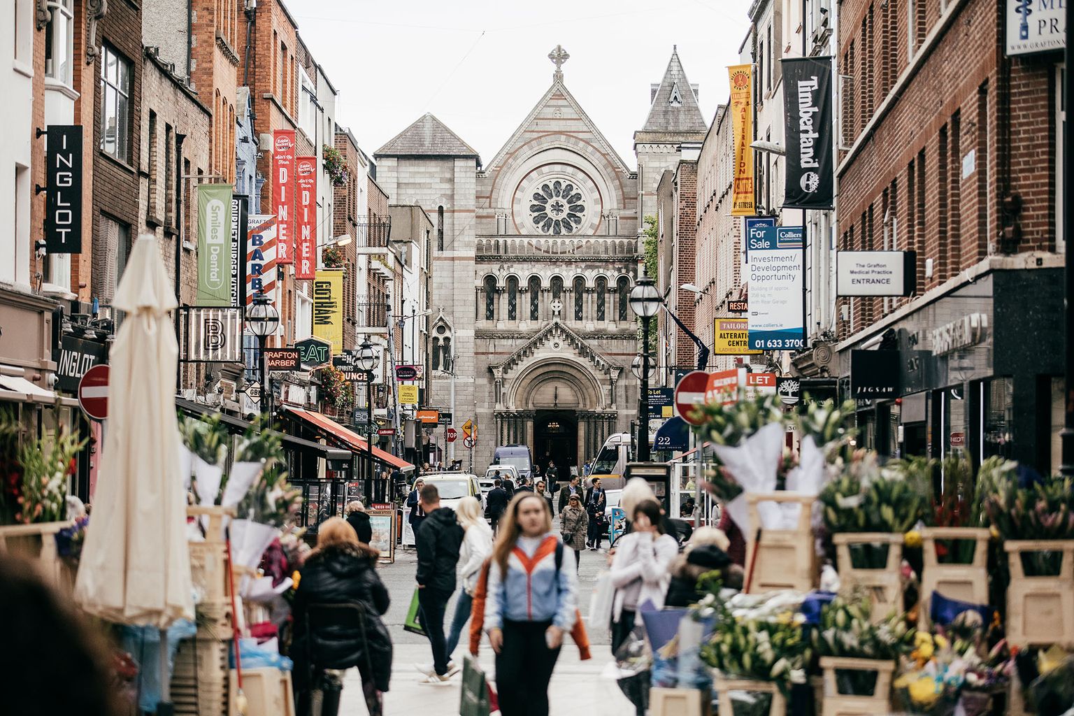 Busy city street with shops and flower stalls, leading to a historic stone church with a rose window and arched entrance in the background.
