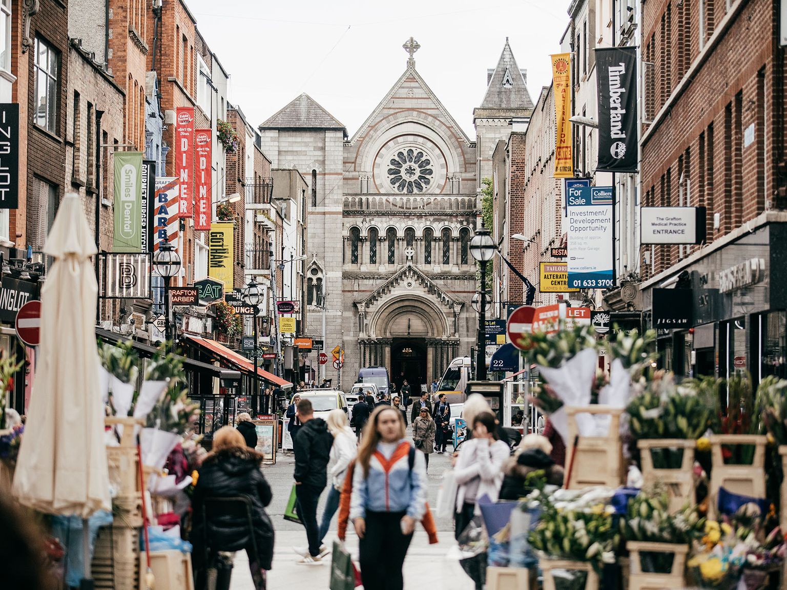 Busy city street with shops and flower stalls, leading to a historic stone church with a rose window and arched entrance in the background.