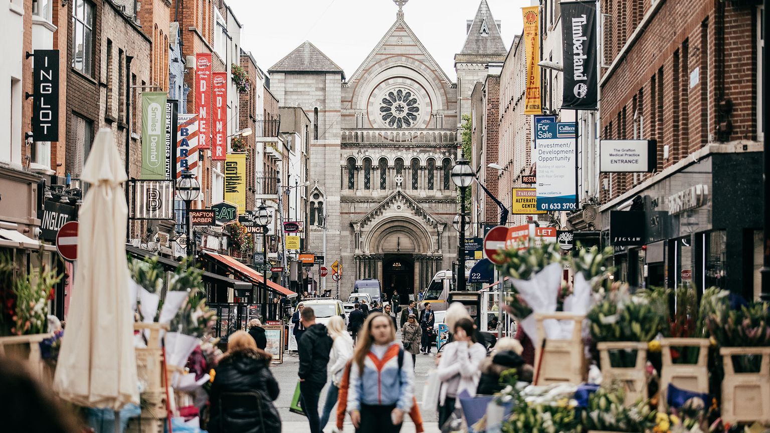 Busy city street with shops and flower stalls, leading to a historic stone church with a rose window and arched entrance in the background.