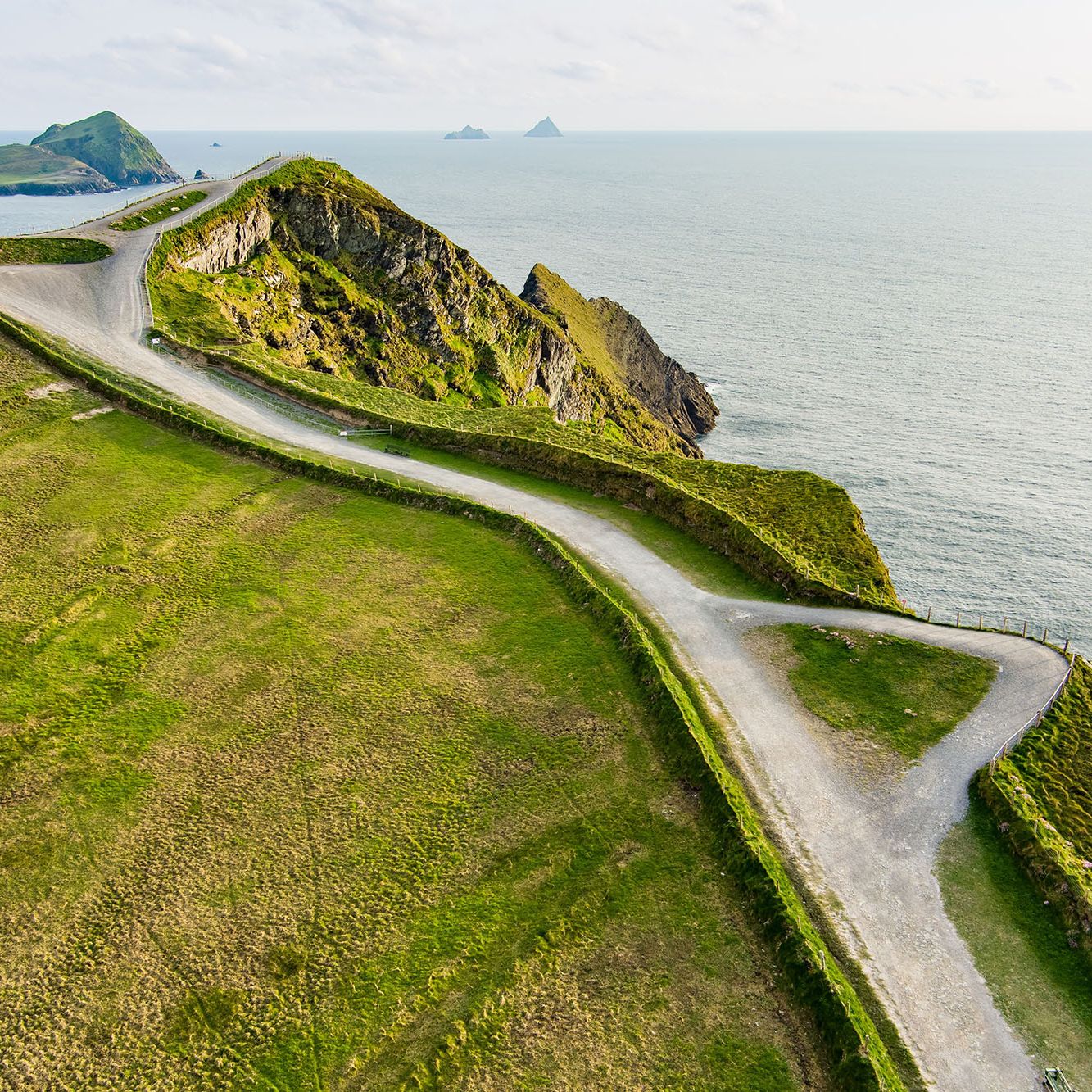 A winding coastal path atop lush green cliffs overlooking a calm sea, with distant islands visible on the horizon.
