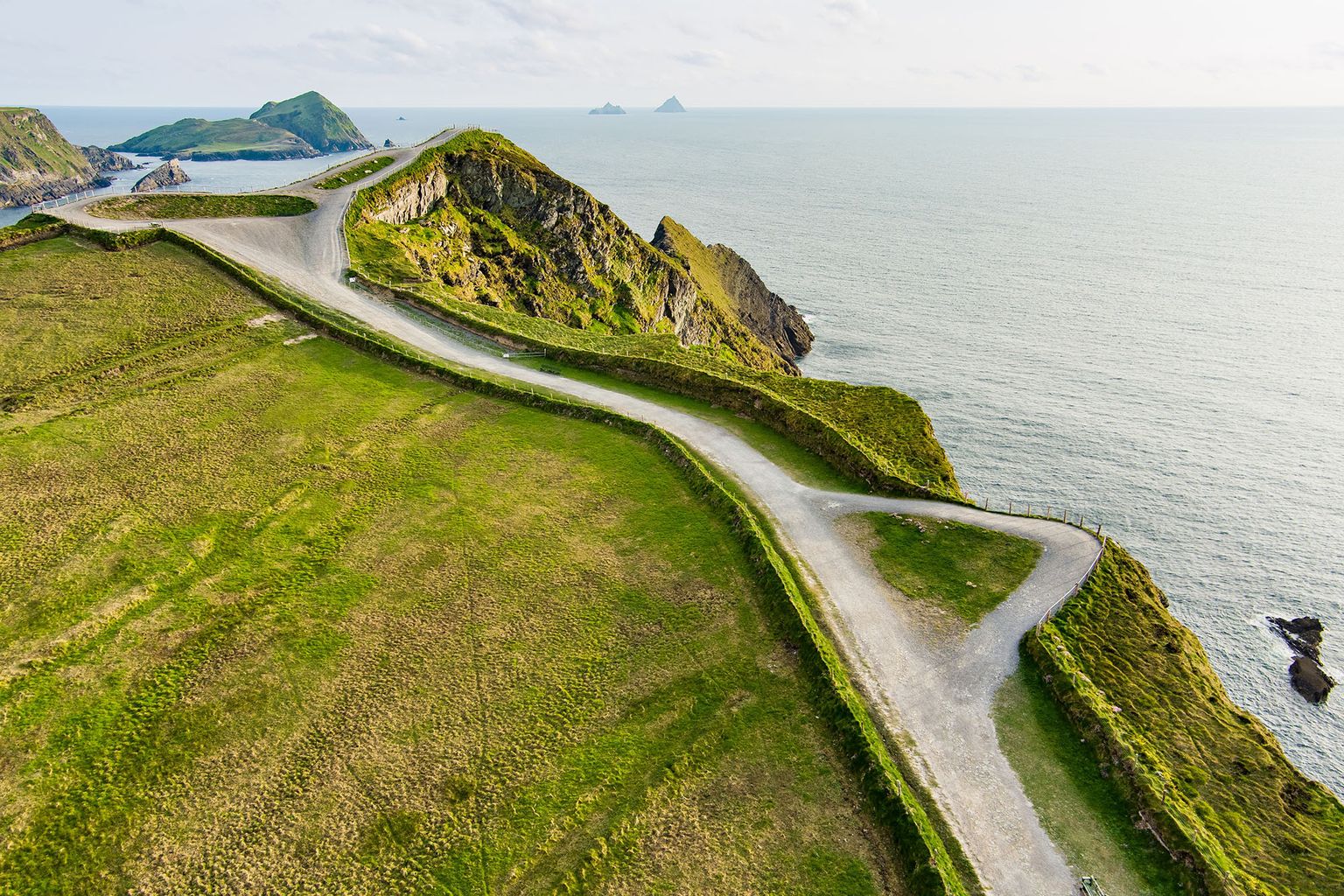 A winding coastal path atop lush green cliffs overlooking a calm sea, with distant islands visible on the horizon.