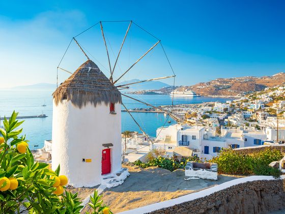 A traditional white windmill with a thatched roof overlooks a coastal village in Mykonos, Greece. Bright yellow lemons adorn nearby foliage.