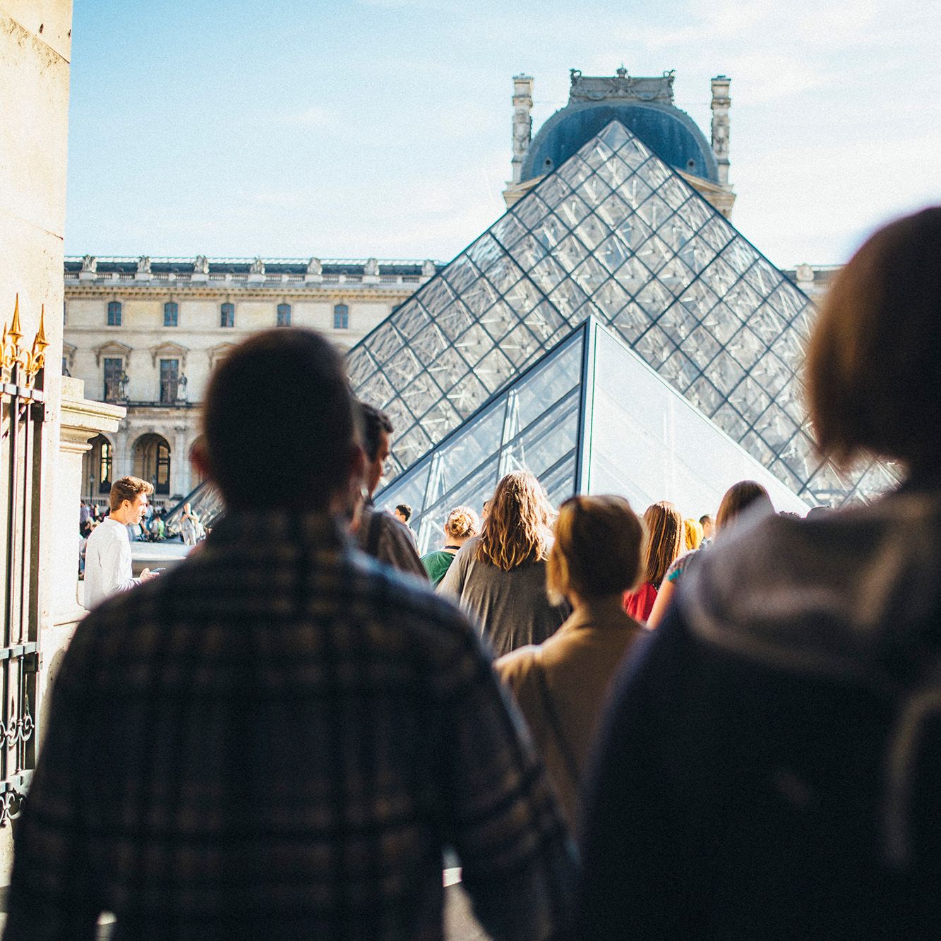 Visitors walk towards the glass Pyramid entrance of the Louvre Museum, with historic buildings in the background under a clear sky.