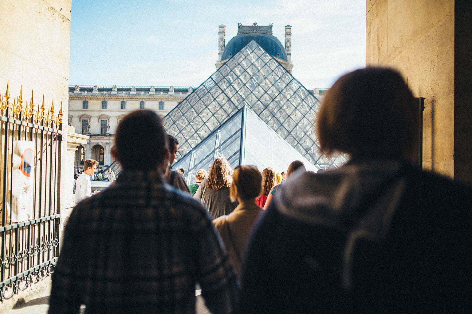 Visitors walk towards the glass Pyramid entrance of the Louvre Museum, with historic buildings in the background under a clear sky.