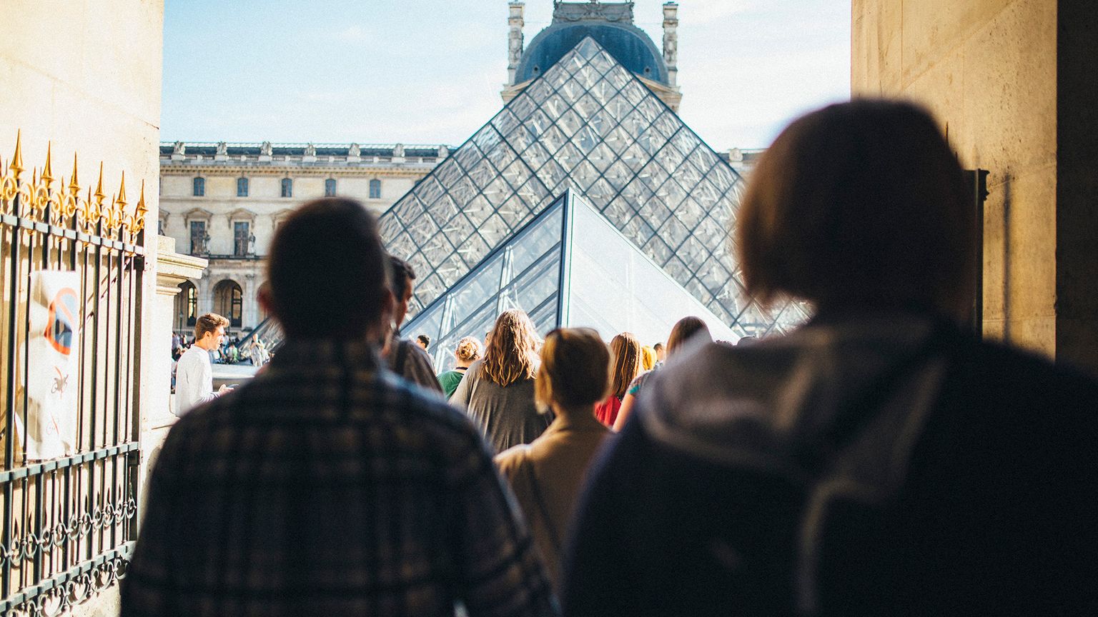 Visitors walk towards the glass Pyramid entrance of the Louvre Museum, with historic buildings in the background under a clear sky.