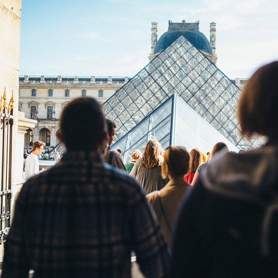 Visitors walk towards the glass Pyramid entrance of the Louvre Museum, with historic buildings in the background under a clear sky.