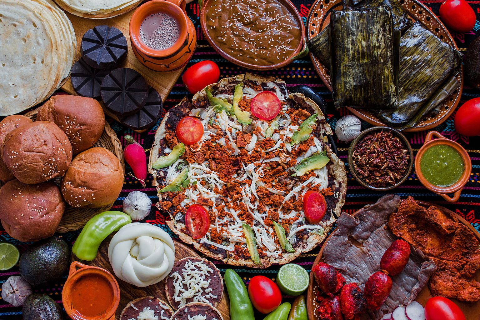 An assortment of traditional Mexican foods including tortillas, bread, tamales, mole, and garnished with fresh vegetables and sauces.