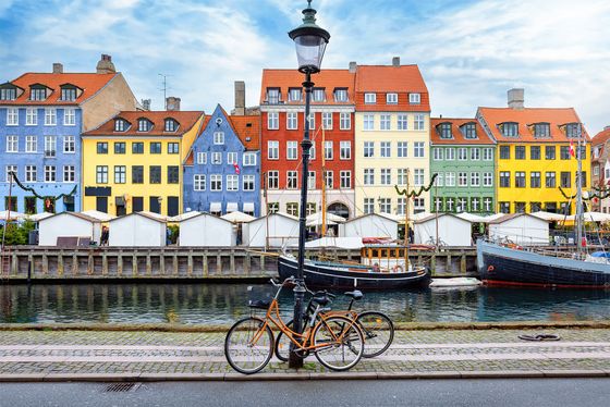Bicycles parked by a lamppost in front of colorful buildings and boats along a canal in a scenic urban setting.