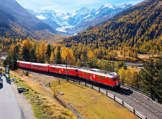 Red train travels through an autumnal mountain landscape with snow-capped peaks in the background.