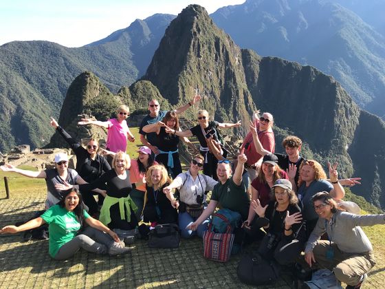 A group of people joyfully pose with arms raised in front of Machu Picchu, with mountains in the background under a clear blue sky.