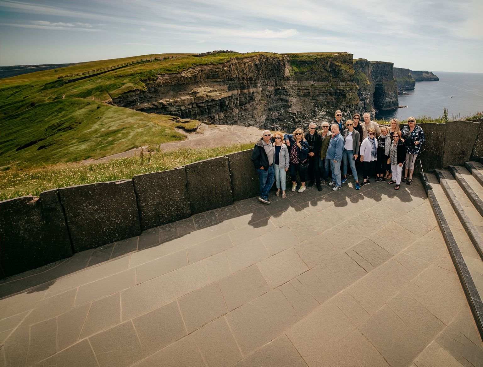 A group of people posing on a viewing platform in front of the Cliffs of Moher, with grassy cliffs and ocean in the background.