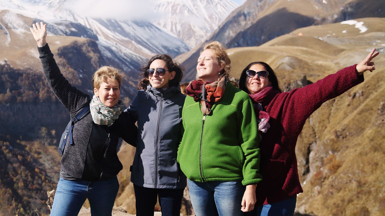 Four people joyfully pose with arms raised against a mountainous backdrop, dressed in colorful jackets, under a partly cloudy sky.