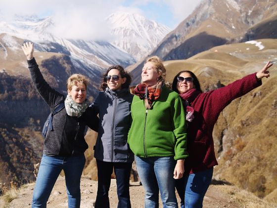 Four people joyfully pose with arms raised against a mountainous backdrop, dressed in colorful jackets, under a partly cloudy sky.