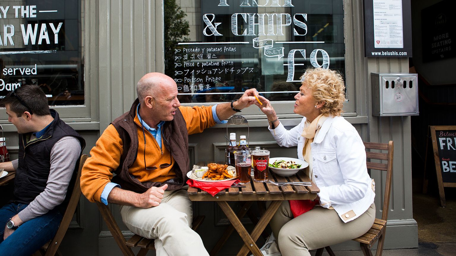 A man and woman smiling, sitting at a cafe table outdoors, sharing a fist bump, with fish and chips on the table, next to a window sign.