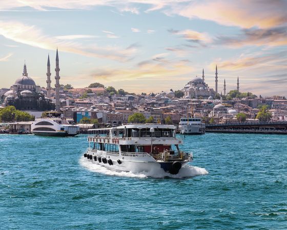 A ferry crosses the blue waters of Istanbul with the city's skyline and mosques under a colorful sky in the background.