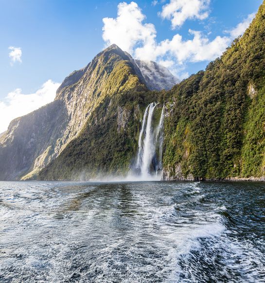 A majestic waterfall cascades down a lush, green mountain into a clear body of water, under a bright blue sky with scattered clouds.
