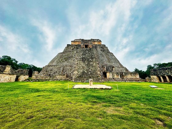 Pyramid with steep stone steps, surrounded by trees under a cloudy sky, on a grassy field.