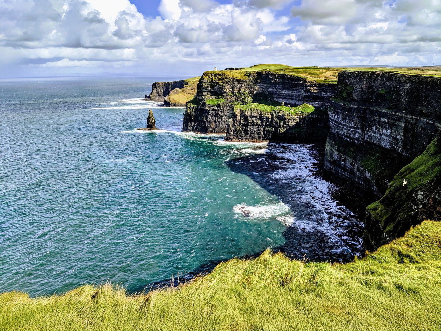 Cliffs overlooking blue ocean under a partly cloudy sky, with green grassy terrain on top and waves crashing below.