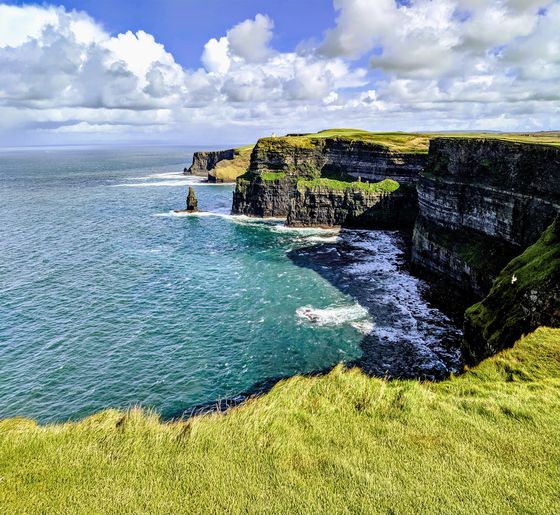 Cliffs overlooking blue ocean under a partly cloudy sky, with green grassy terrain on top and waves crashing below.
