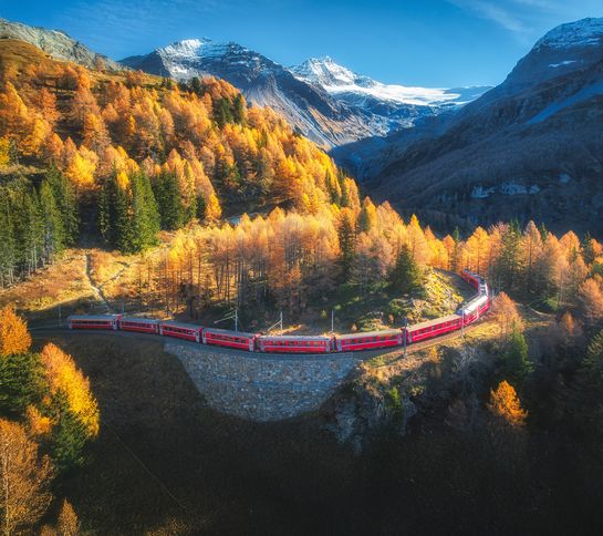 Red train travels through vibrant autumn forest with snowy mountain backdrop under clear blue sky.