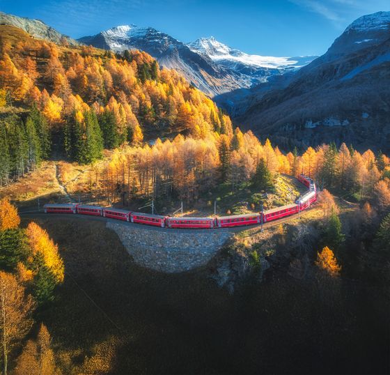 Red train travels through vibrant autumn forest with snowy mountain backdrop under clear blue sky.