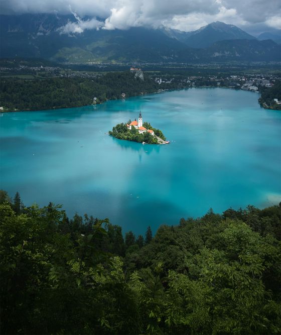 Aerial view of a turquoise lake with a small island featuring a church, surrounded by lush forests and mountains under cloudy skies.
