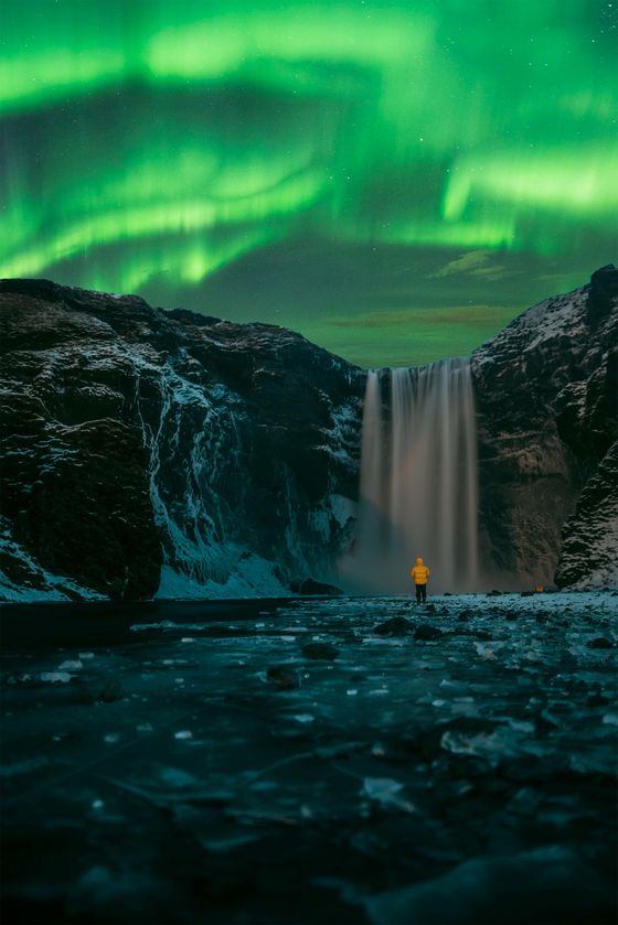 Person in a yellow coat stands before a waterfall under vibrant green Northern Lights, surrounded by snowy landscape.