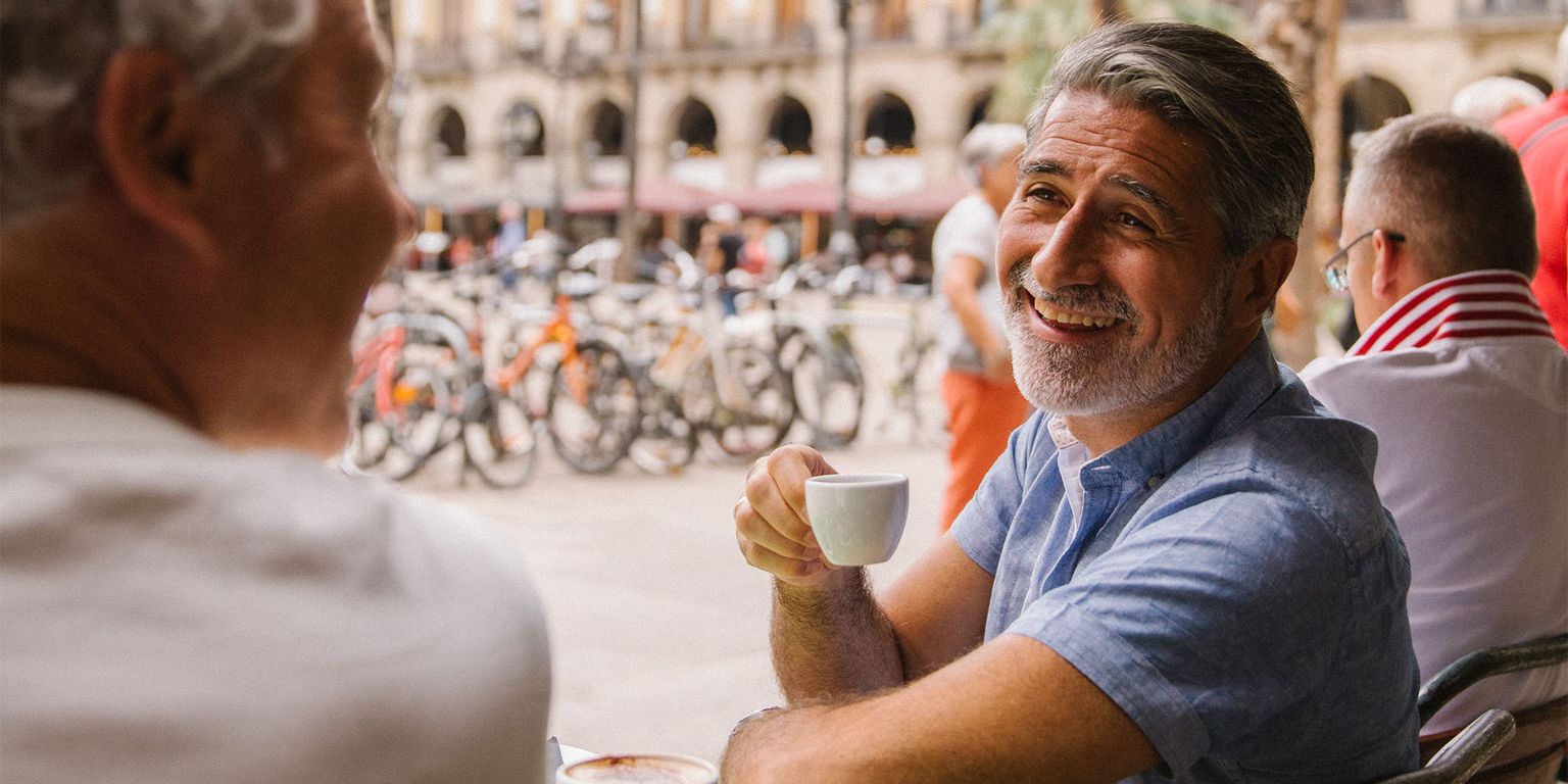 Smiling man in a blue shirt enjoys coffee at an outdoor café, surrounded by palm trees and bicycles, talking to a friend.