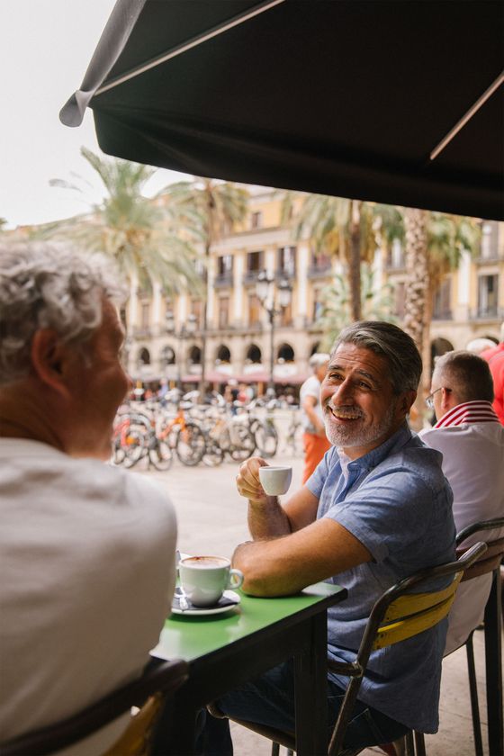 Smiling man in a blue shirt enjoys coffee at an outdoor café, surrounded by palm trees and bicycles, talking to a friend.