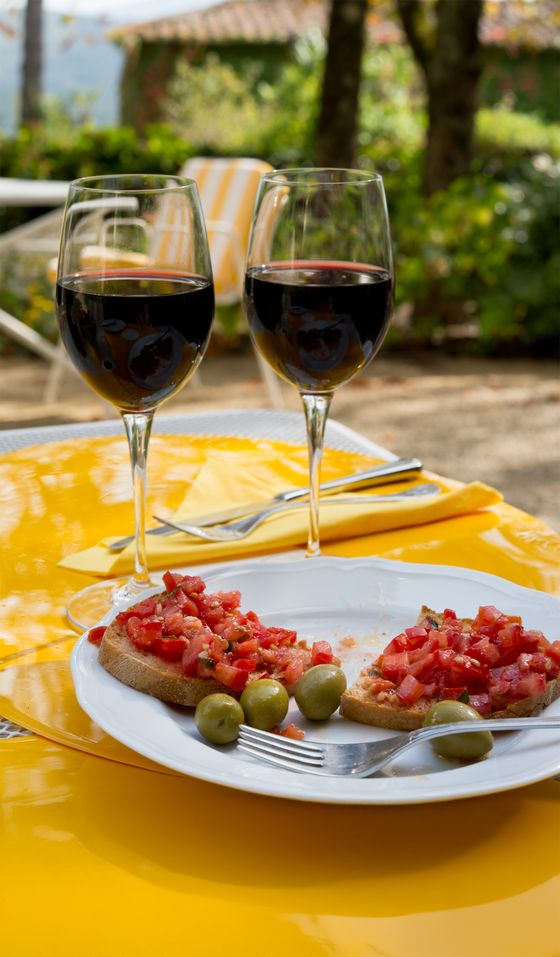 Two glasses of red wine and bruschetta topped with tomatoes and olives on a yellow outdoor table setting.