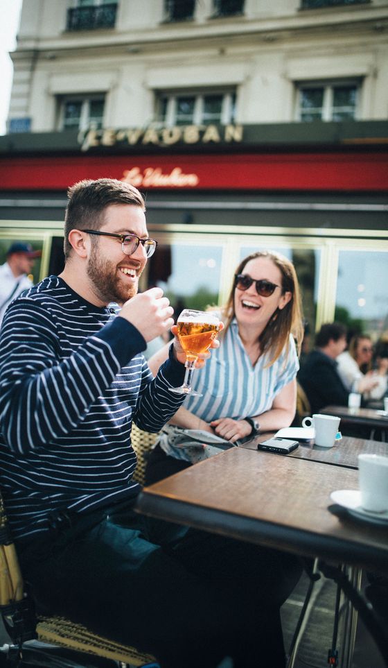 Two people laughing and enjoying drinks at an outdoor café. One holds a glass of beer, the other has sunglasses on. Sunny day with a relaxed atmosphere.