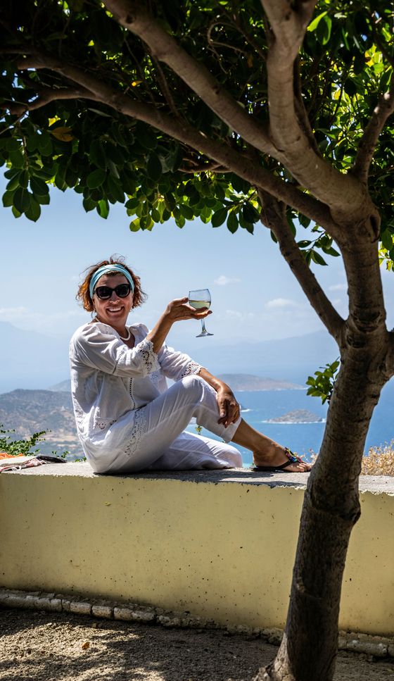 Smiling person in white outfit sitting on a ledge, holding a wine glass, with a scenic view of the sea and mountains in the background.