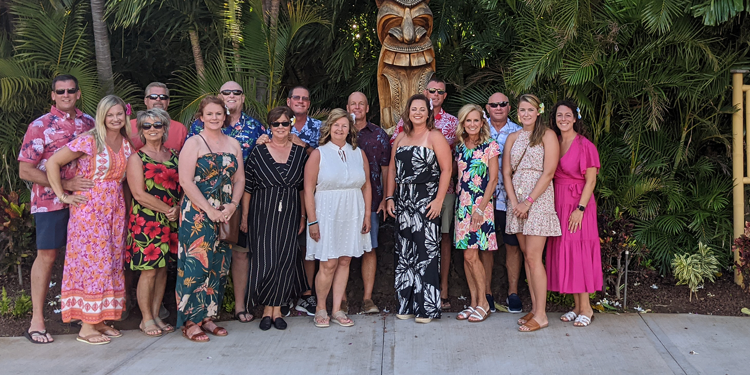 A group of people in colorful attire pose together outdoors near a "Luau" sign surrounded by lush greenery and a wooden statue.