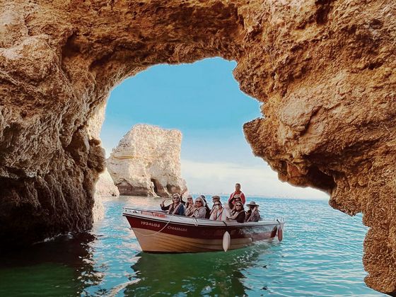 A small boat with people navigates through a rocky sea cave, turquoise water below and blue sky above.