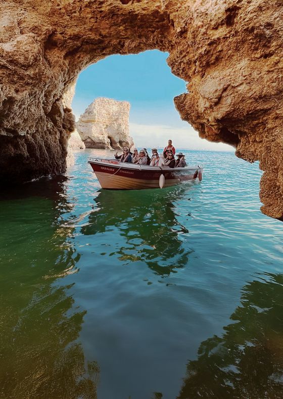 A small boat with people navigates through a rocky sea cave, turquoise water below and blue sky above.