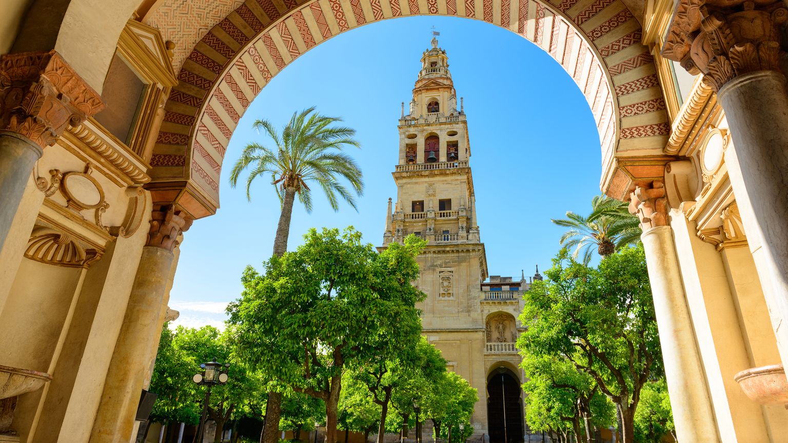 The Great Mosque of Córdoba's bell tower framed by an ornate arch, surrounded by lush orange trees and vibrant blue sky.