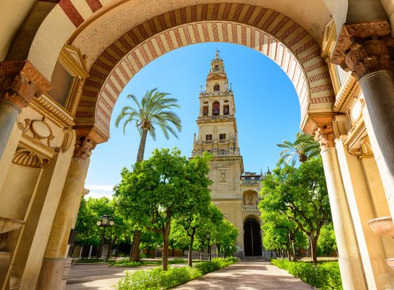 The Great Mosque of Córdoba's bell tower framed by an ornate arch, surrounded by lush orange trees and vibrant blue sky.
