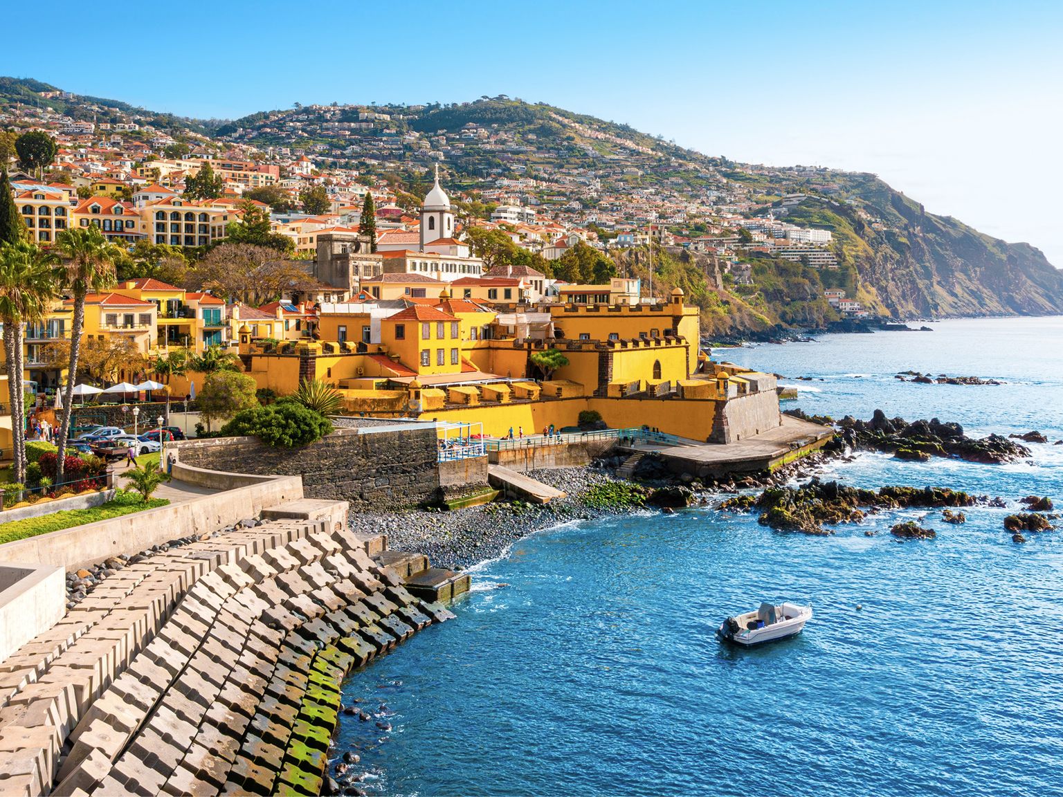 Coastal town with vibrant buildings, a church tower, a boat on the water, rocky shoreline, and hillside houses under a clear blue sky.