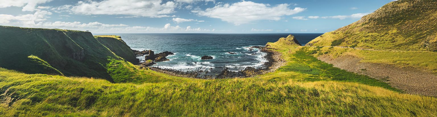 Coastal landscape with grassy cliffs framing a rocky shoreline, under a partly cloudy sky, and blue ocean in the background.