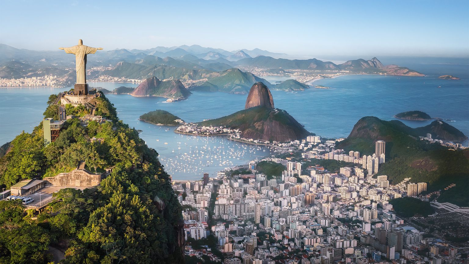 Aerial view of Rio de Janeiro featuring Christ the Redeemer, Sugarloaf Mountain, and the sprawling cityscape against a blue ocean backdrop.